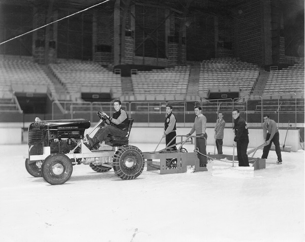A young man on a tractor drives on an ice arena with a trailing plow. 5 others stand behind holding shop mops