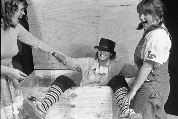 Two women trying to help a third woman out of a large wooden box.