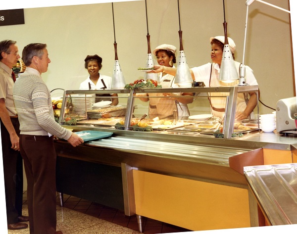 Cafeteria workers handing food to two men holding lunch trays