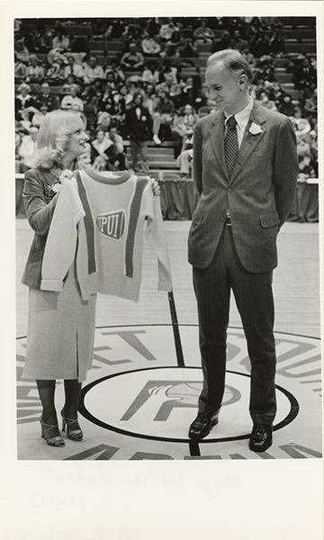Tall man in a suit standing on the center line of a basketball court with a woman presenting him with a long-sleeve IUPUI sweater
