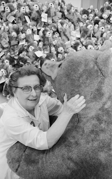Older woman holds. a giant teddy bear, with many small teddy bears in the background