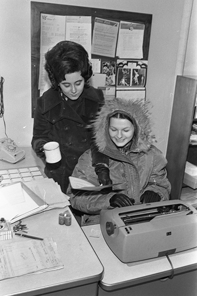 Two women in coats in a standard office working on a typewriter