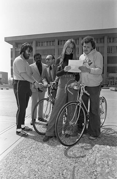 Two young people stand next to thier bikes while holding a paper between them. three other people stand behind them
