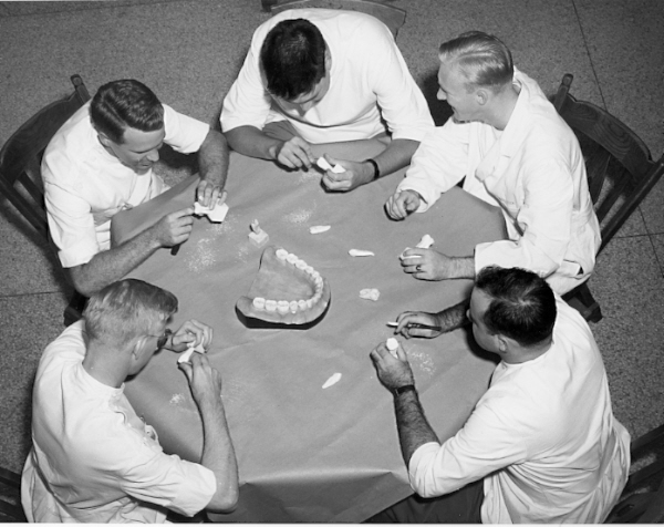A high shot looking down at five men gathered around a circle table carving teeth. A giant teeth model sits in the middle of the table.