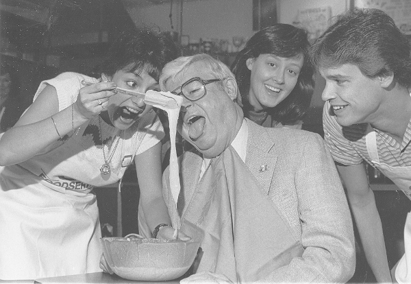 A man has his mouth open while an enthusiastic woman feeds him pancake batter. Two young students look on with smiles
