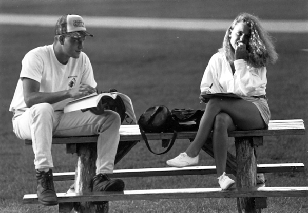 A young man in a baseball uniform is looking at a big open book while a woman looks away from her writing. They are seated on the table of a park bench