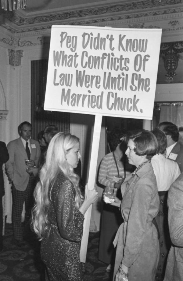 Two women staring at one another. One is holding a sign that says, 'Peg didn't know what conflicts of law were until she married Chuck"