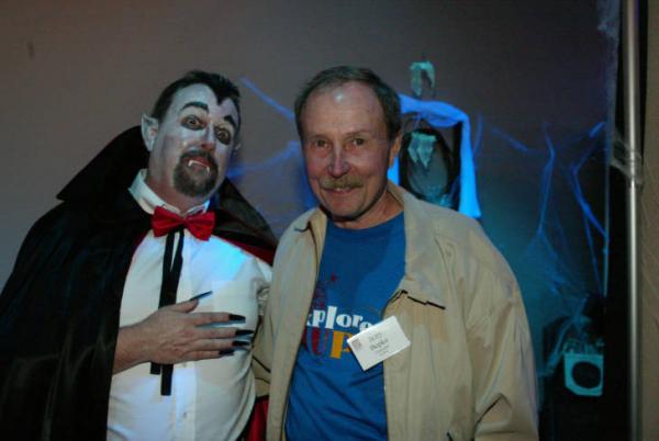 Older man with a mustache stands smiling next to another man dressed as a vampire
