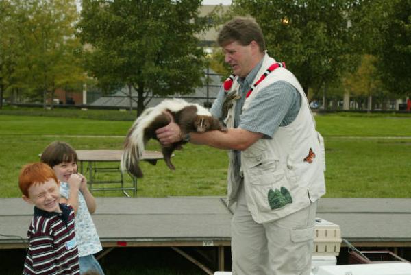 Zoo keeper holds up the tail end of a skunk above two children with disgusted looks on their faces