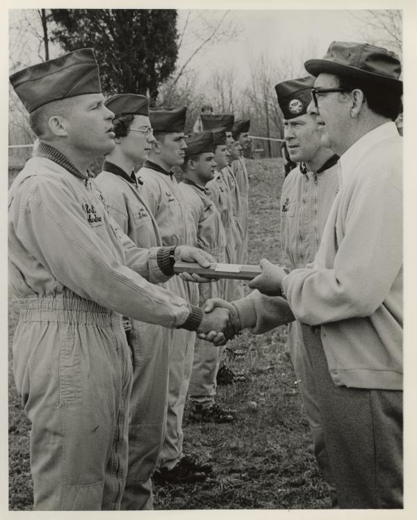 Man in a cardigan and glasses shaking hands with a soldier while they hold a cylinder between them. Other soldiers are lined up beside them