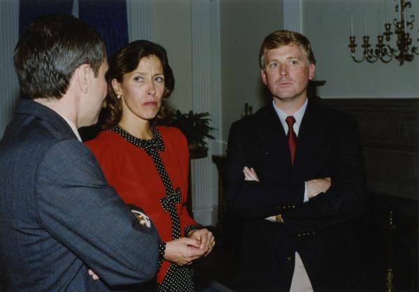 Color photograph. A man in a gray suit is facing away from the camera, talking to a man and a woman. The woman has dark hair and is wearing a red dress with black and white polka dots along the collar and middle. She's looking at the speaking man with an expression of incredulousness. The other man is blonde and is wearing a black suit with a red tie. His arms are crossed and he's looking out of frame. His expression is one of exasperation.