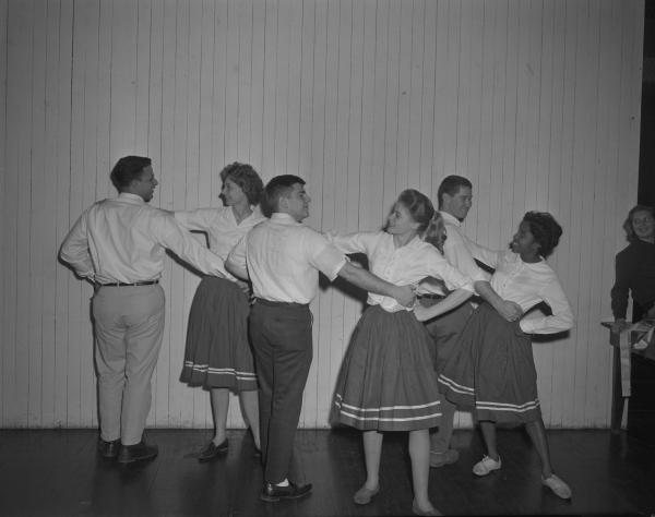 Black and white photograph. Three couples are dancing together, with the men on the left of their partners and the women on the right. The women have a hand on their partner's shoulder and the other in a fist on their hips. The men are posed similarly, but with their hand on their partner's waist. Everyone is wearing a button up shirt, with the women wearing skirts with two horizontal white lines near the bottom. On the right hand side of the picture a smiling woman is leaning on a table watching them dance. On the table is a sash.