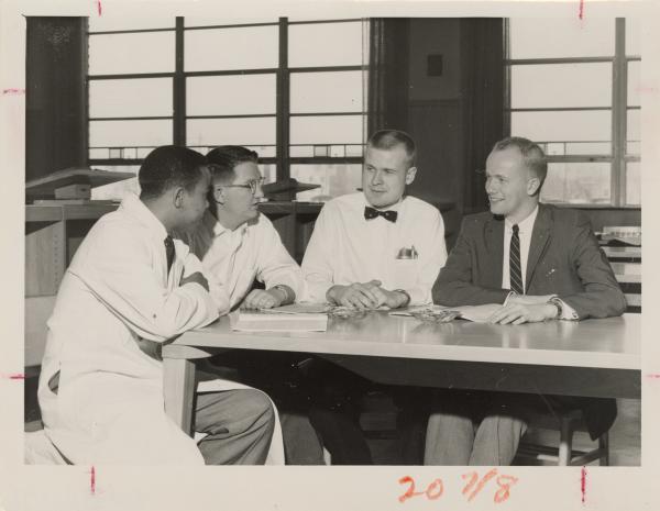 Black and white photograph. Four men (the leftmost is black, while the others are white) sit around a rectangular table looking at each other. The three men on the left hand side of the photo are wearing labcoats, the rightmost is wearing a suit and tie. The man in the middle left is leaning forward as if saying something important. The man to the right of him looks taken aback, but has the hint of a smile on his face. The man in the suit is smiling. On the table sits an open, typed document and an ashtray. Behind the men are large windows looking outside. It appears that they are on the second or third floor. A pair of lecterns are also behind them. At the bottom, in the margins of the photo, 20 7/8 is written in red pencil.