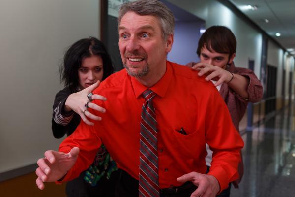 Color photograph. Older, white, man with gray hair and a a gray goatee takes up most of the frame, posed as if he were running or trying to escape.  He wears a bright red dress shirt and a red and gray striped necktie. Pens are visible in his breast pocket. Two younger people (a man and woman) are being him, their hands curled over his shoulders as if to grab him. The woman is wearing three rings on her hand, and has heavy eye-liner and eye shadow. Her hair is short and dark. The young man ears an open dress shirt over a white t-shirt. On his cheek is a black line. The background is a school hallway.