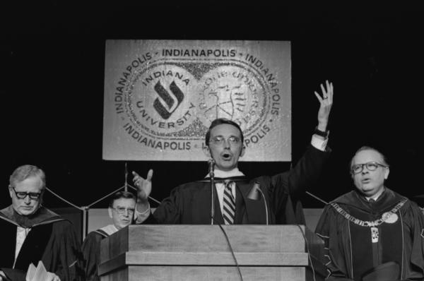 Black and white photograph. A middle aged white man wearing glasses is standing a podium with microphones on it. His arms are raised and his mouth is open, as if he is singing or mid speech. There are three other white man behind him. Everyone is wearing graduation gowns. Behind them it's all black except for a white sign that has the word Indianapolis wrapped around two circular logos, one of IU the other Purdue.