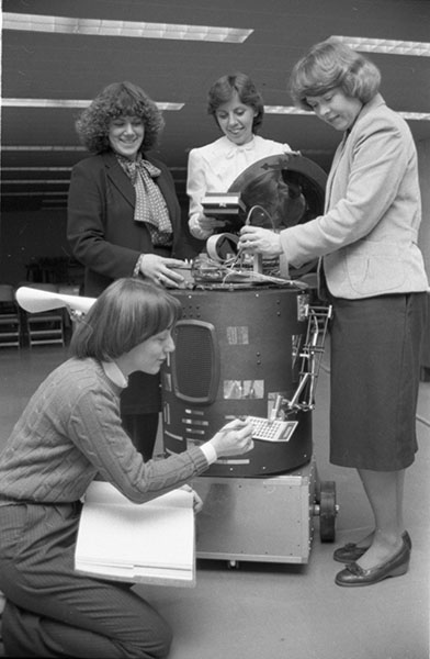 Three woman stand around a cylindrical robot while a fourth kneels in front of it