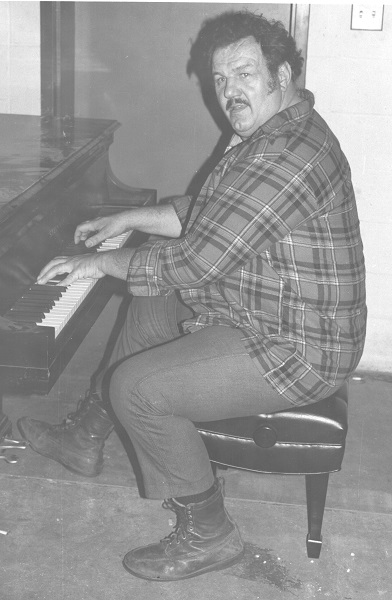 Large man with a mustache looks into the camera while playing piano