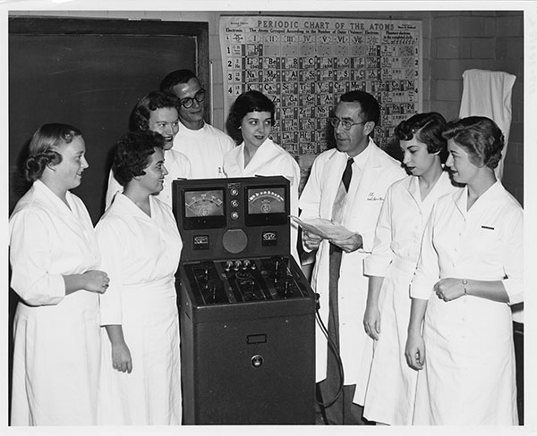 Seven young people and an older man, all in white work clothes, stand around a tall machine