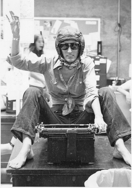 Woman wearing an aviator cap and glasses is seated on a table in front of a typewriting. She is miming that she is controlling the typewriter as a vehicle