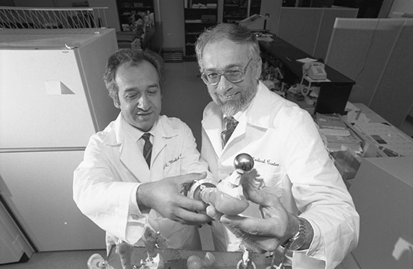 Two older men in lab coats hold a ball joint attached to a fake bone to the camera