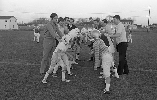 Football teams of very young boys face eachother on the field, while adult man stand behind them pressing their shoulders