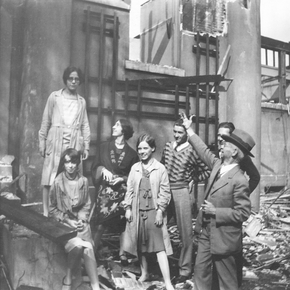 Seven people standing in the ruins of a building. Theman in the foreground has his arm extended upwards, showcasing the damage