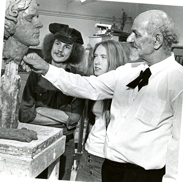 Old balding man pressing clay into a bust while two students watch