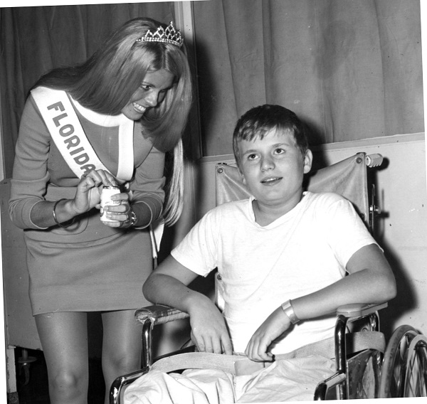 Woman in a tiara and sash reading, 'Florida,' stands over a young boy seated in a wheelchair