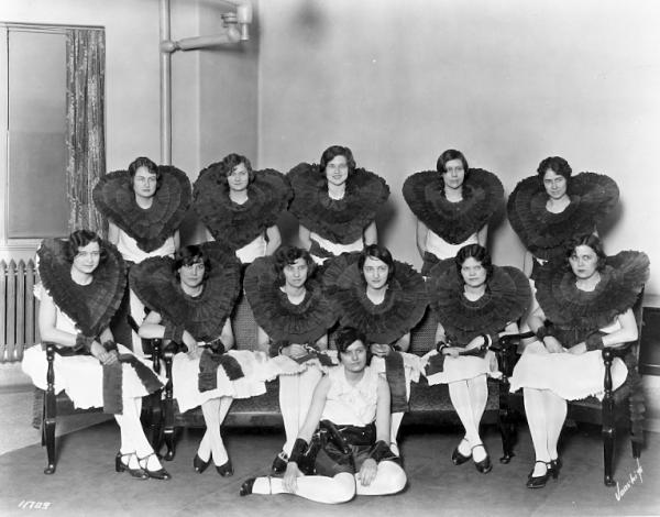 12 women sit posed for a photo. One is on the floor and in a sleeveless dress shirt. The others are seated in chairs, wearing big ruffled neck collars