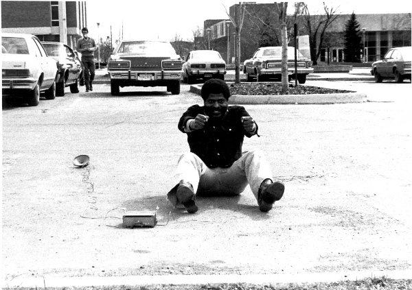 A man is seated in a parking lot, in a position to mime driving, with a receiver and speaker on the ground next to him