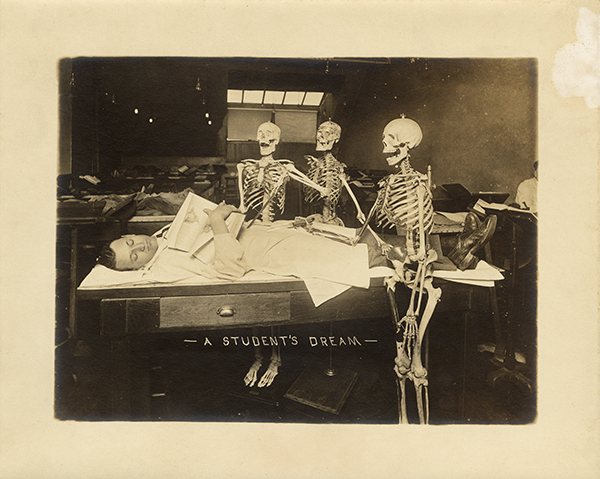 Three skeletons posed around a student laying on a desk asleep. A caption is scratched into the photo: A Student's Dream