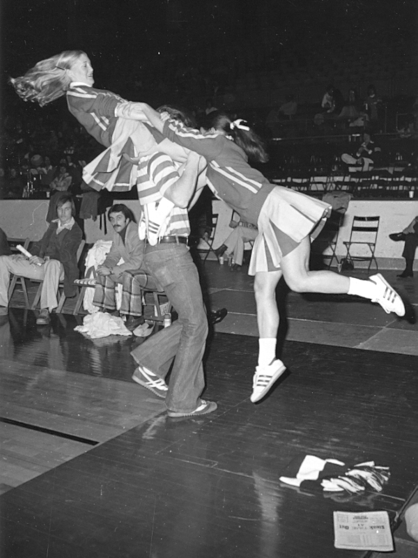 A female cheerleader seated on a male cheerleader's shoulders hold hands with another female cheerleader as they all spin around. 
