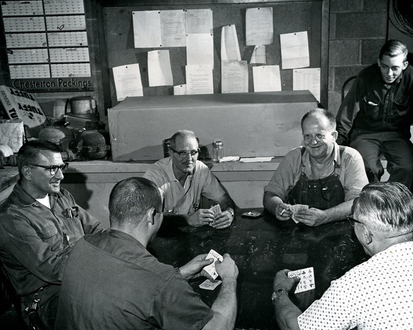 Five older men sit around a table playing cards with a young man sits away watching