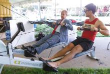 An older man in a suit and tie uses a rowing machine next to a younger man in shorts and an IUPUI tanktop