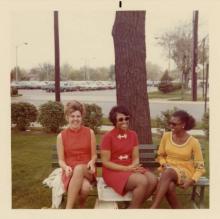 Three young women, one white and two black, sit on a bench. Behind them is a large tree and a parking lot