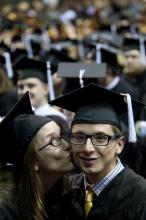 A young woman kisses the cheek of a young man. Both are wearing graduation outfits