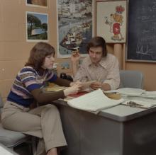 A man and woman are sititng at a desk. The woman is looking at a piece of paper while the man looks at her intently. He has a cigarette in his hand