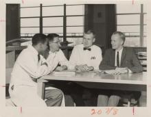 Four men (the leftmost is black, while the others are white) sit around a rectangular table looking at each other. The three men on the left hand side of the photo are wearing labcoats, the rightmost is wearing a suit and tie. The man in the middle left is leaning forward as if saying something important. The man to the right of him looks taken aback, but has the hint of a smile on his face. The man in the suit is smiling. On the table sits an open, typed document and an ashtray.