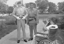 Black and white photograph. The year is 1953. The setting is outdoors on a sidewalk with a manicured lawn in the background. There are trees in the distance and cars from that era parked to the side. A white boy in a wheel chair appears in the foreground. His back is to the photographer, and he holds a camera to his eye. Two men dressed as the Lone Ranger and Tonto are standing in front of the boy. The Lone Ranger is looking skyward above the boy's head. Tonto is staring at the boy's camera.