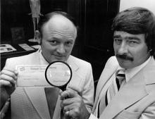 Two men in suits holding a check with a magnifying glass held up to it. They are looking into the camera