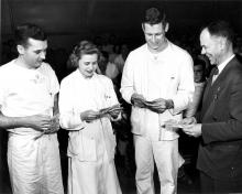Three young people smile while looking at small pieces of paper being distributed by a laughing man in a dark suit