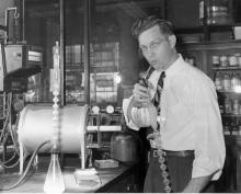 Black and white photograph. The setting appears to be a chemical laboratory from the 1940s or 1950s. There are flasks, jars, vials, and machinery in the room consistent with a lab from that era. A white man wearing glasses, rolled up sleeves, a tie, and gray pants is staring at the photographer. His hair rises oddly on the right side. He has a straw in his mouth which is connected to a lab bottle with a long circular tube dangling towards the floor.