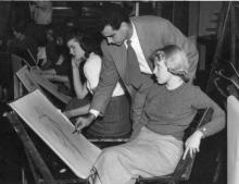 Man in a tie leans over a young woman, demonstrating figure drawing. the woman is focused on the model (who is off camera)