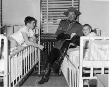 Black and white photograph. A smiling white man with mustache in a Canadian Yukon uniform holds an open book while sitting on a hospital bed next to a white boy lying down. They are both looking at the camera. The man is smiling and the boy has a serious look on his face. A second white boy in another bed leans over in interest.   The head of the beds are against a wall on opposite sides of a window with closed blinds.
